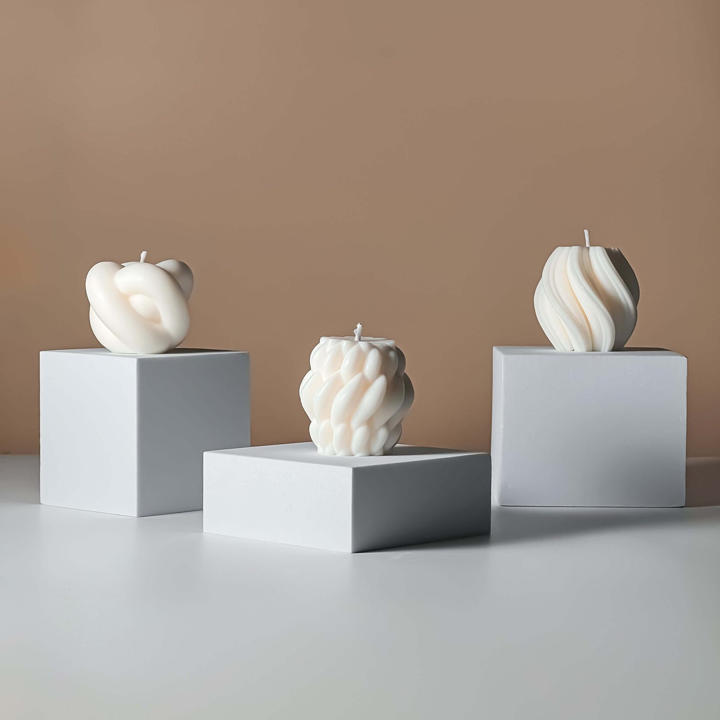 Three abstract candle sculptures on white cubes against a beige wall.