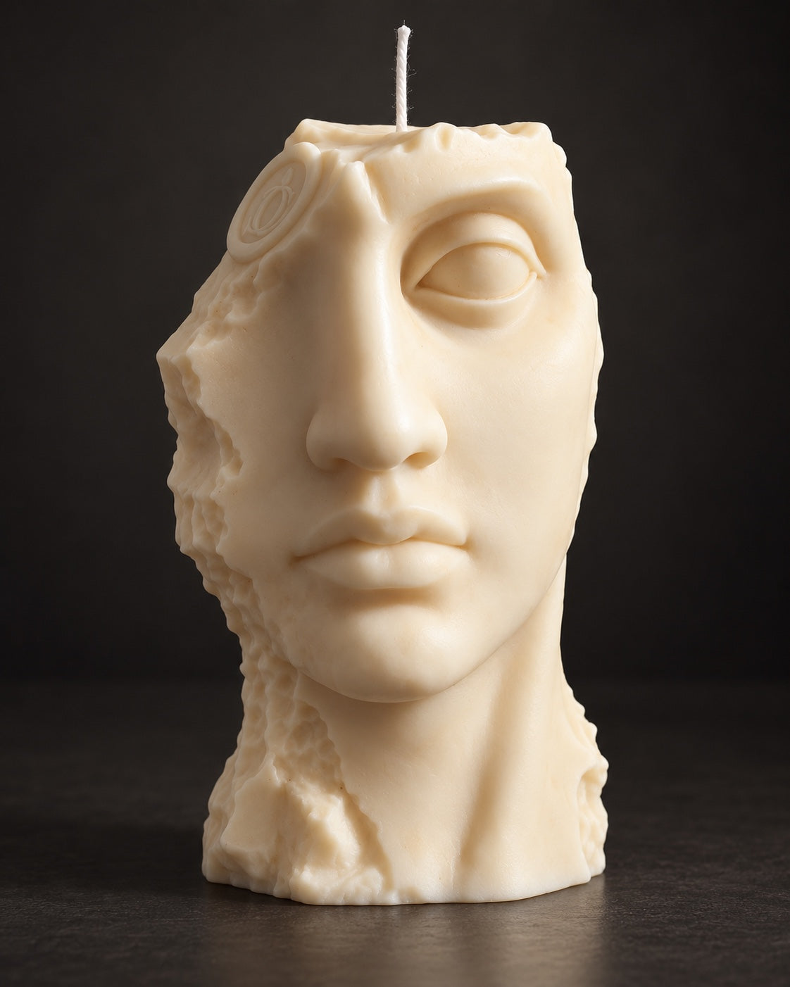 A sculptural candle shaped like a classical female bust head, made of cream-colored wax with a single white wick at the top, set against a dark studio background.