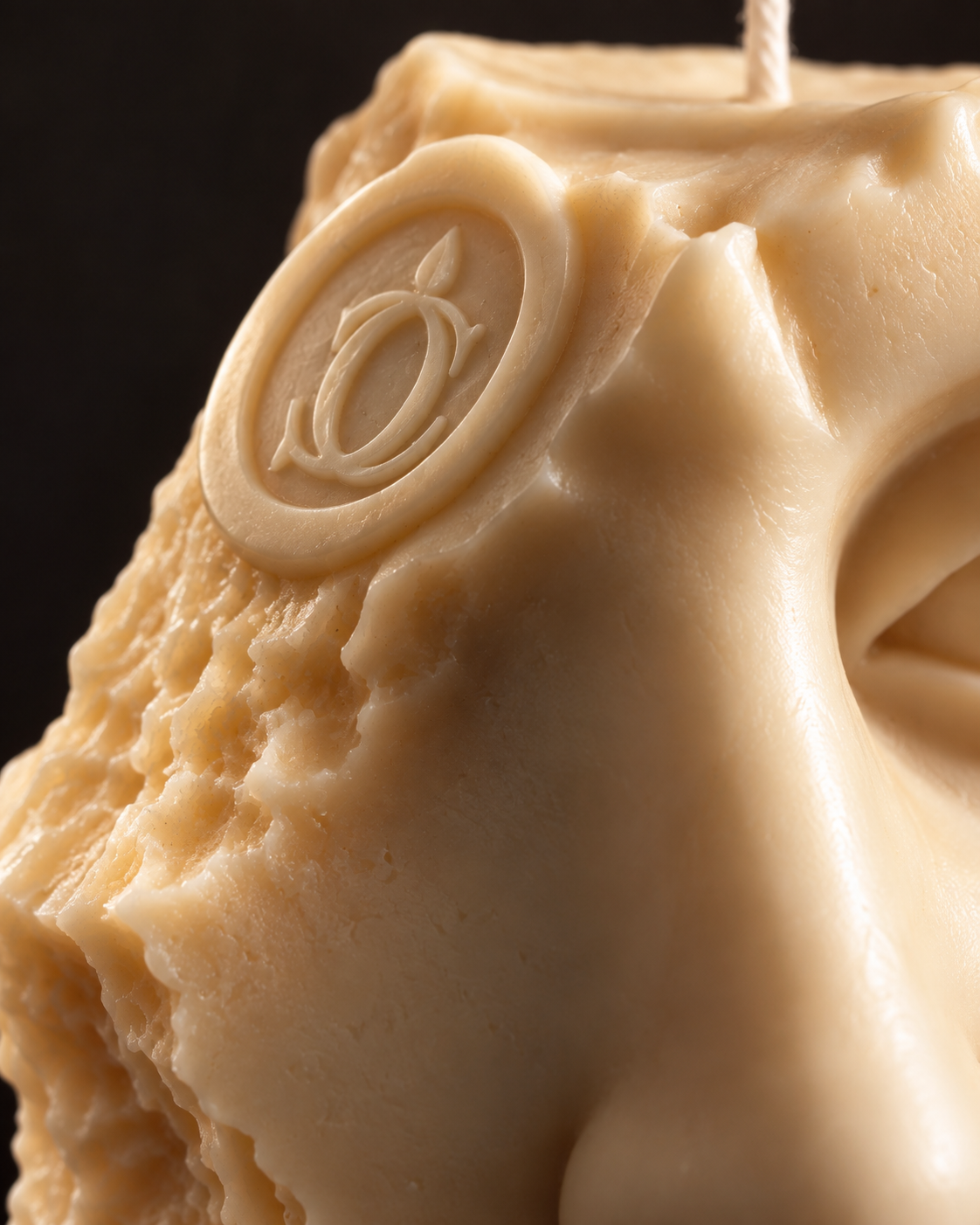 Close-up of a sculpted beige wax candle with a carved circular emblem and textured surface, photographed against a dark background.
