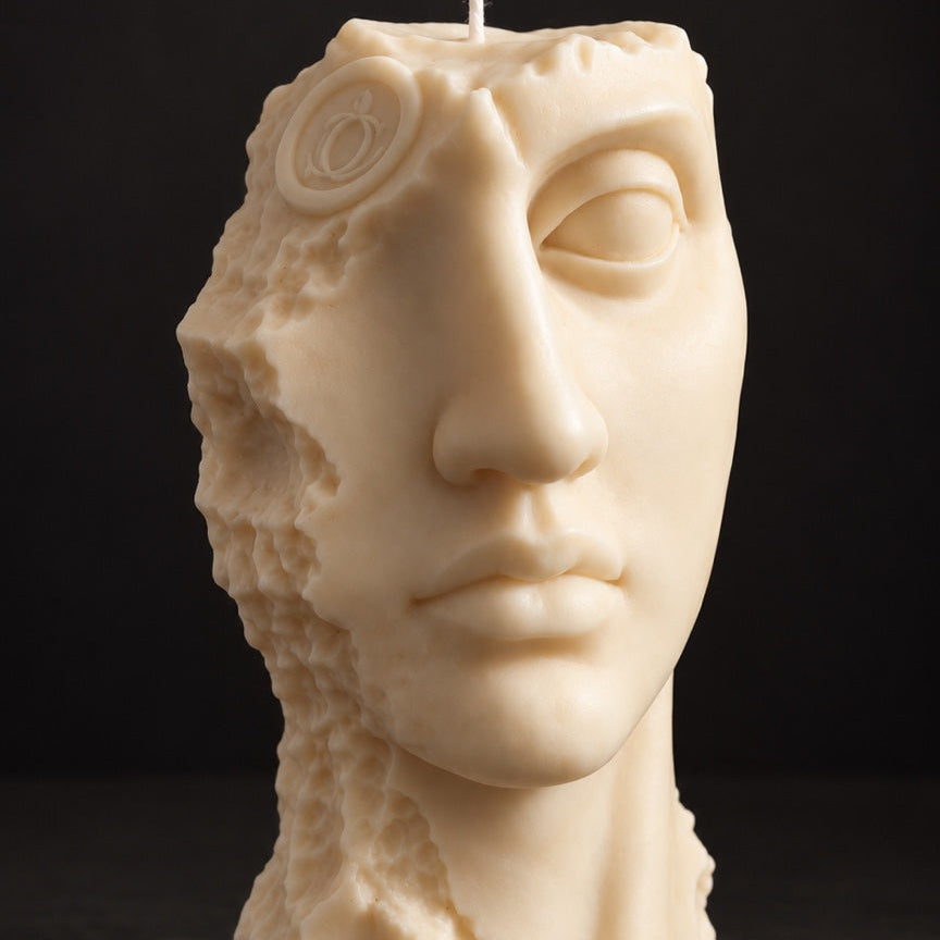 An off-white sculptural candle shaped like a classical human face, with a single wick at the top, photographed against a dark black background.