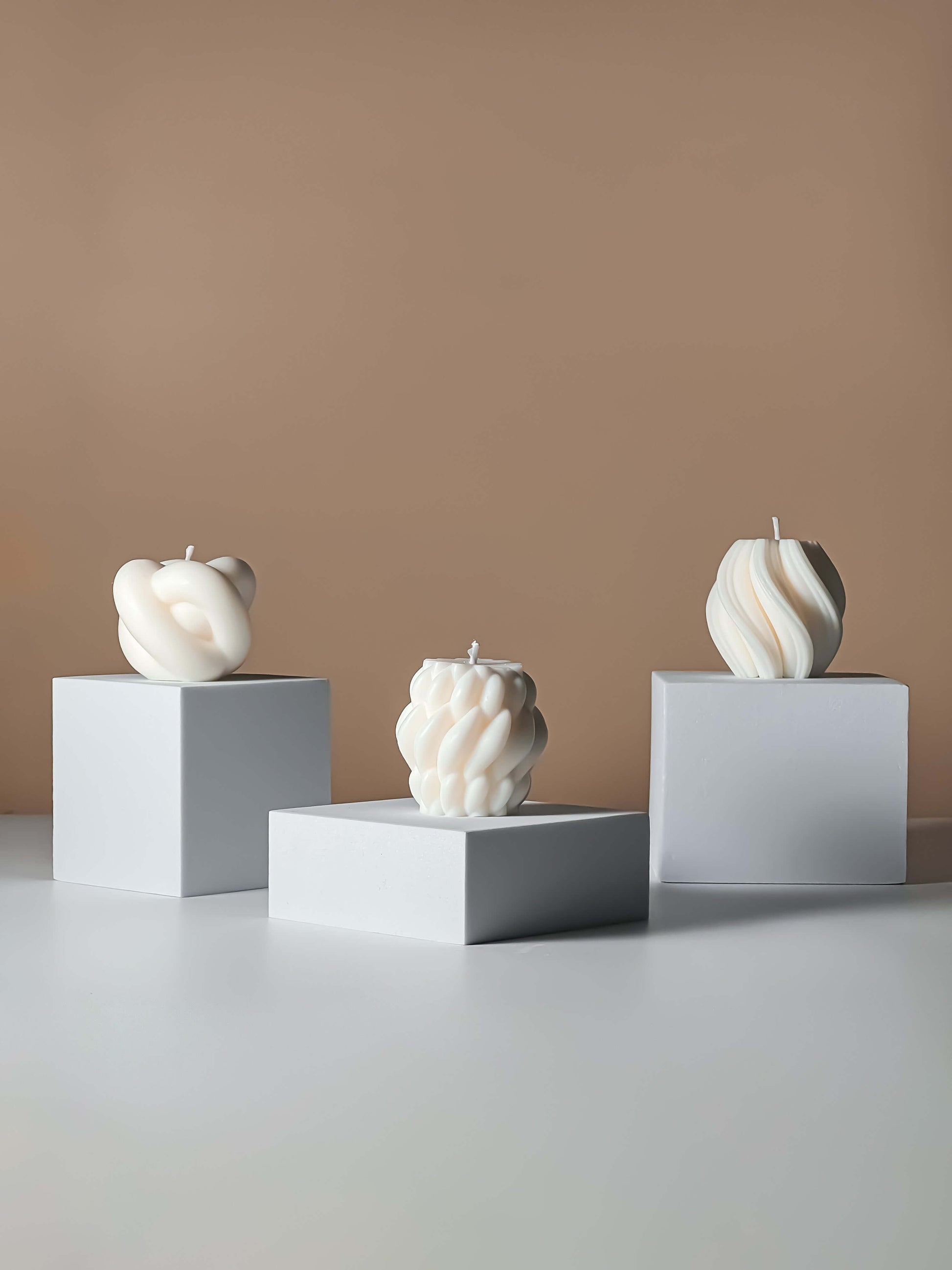Three abstract candle sculptures on white cubes against a beige wall.